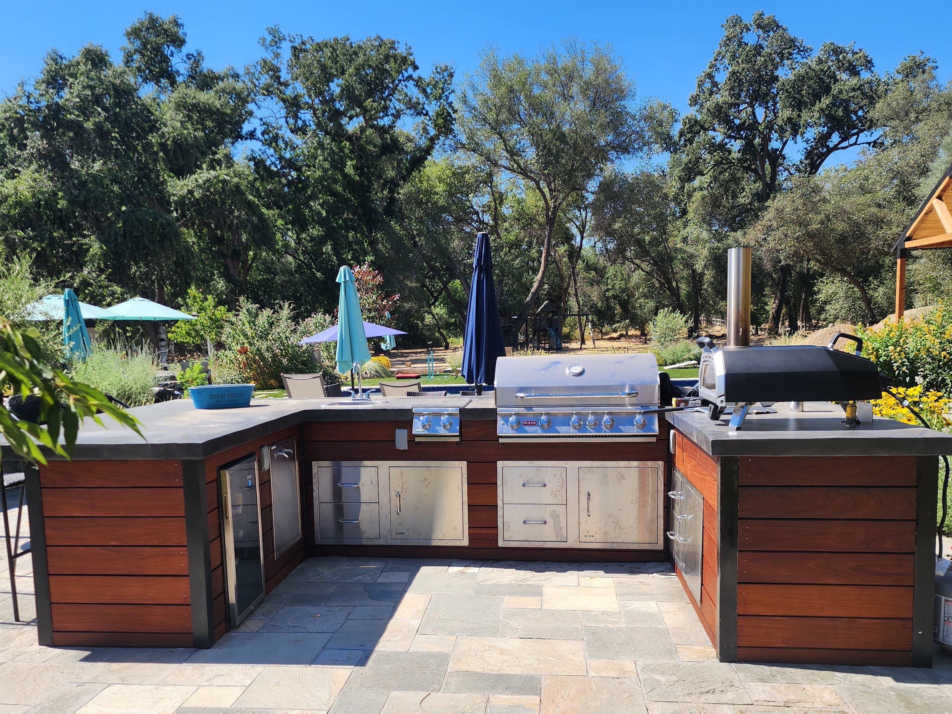 L-shaped outdoor kitchen island with ipe wood cladding, concrete counters, a built-in grill, and a pizza oven
