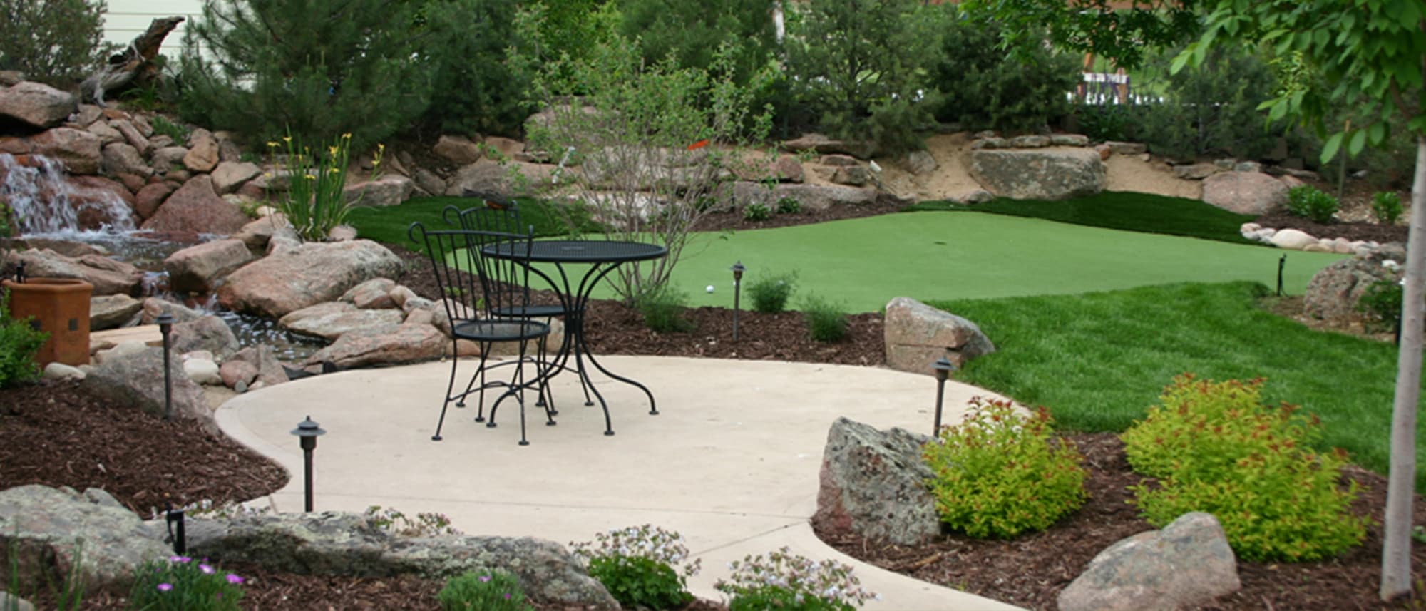 Concrete patio beside a boulder waterfall with an artificial turf putting green beyond