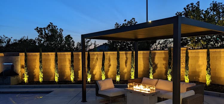 Landscape lighting at dusk uplighting cypress along a wall beside a modern pergola and fire table